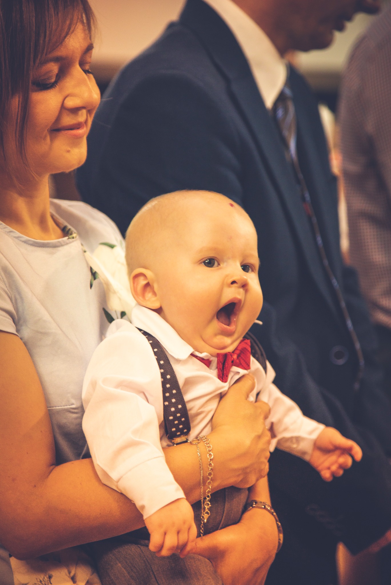 Uśmiechnięta kobieta trzyma na rękach małego chłopca w białej koszuli, czerwonej muszce i szelkach w kropki, który szeroko otwiera buzię, w tle mężczyzna w garniturze