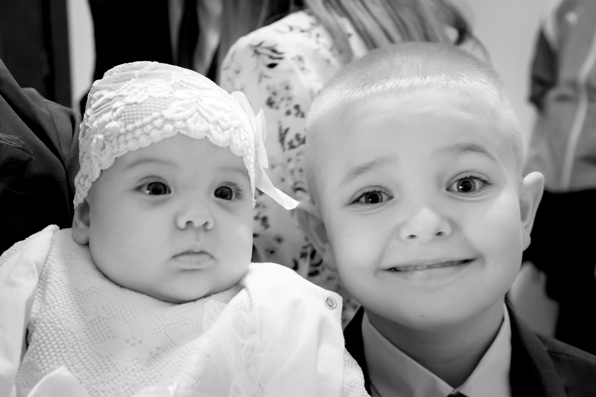 Czarno-białe zdjęcie portretowe niemowlęcia w koronkowej czapeczce i starszego chłopca w garniturze, oboje patrzą w obiektyw.