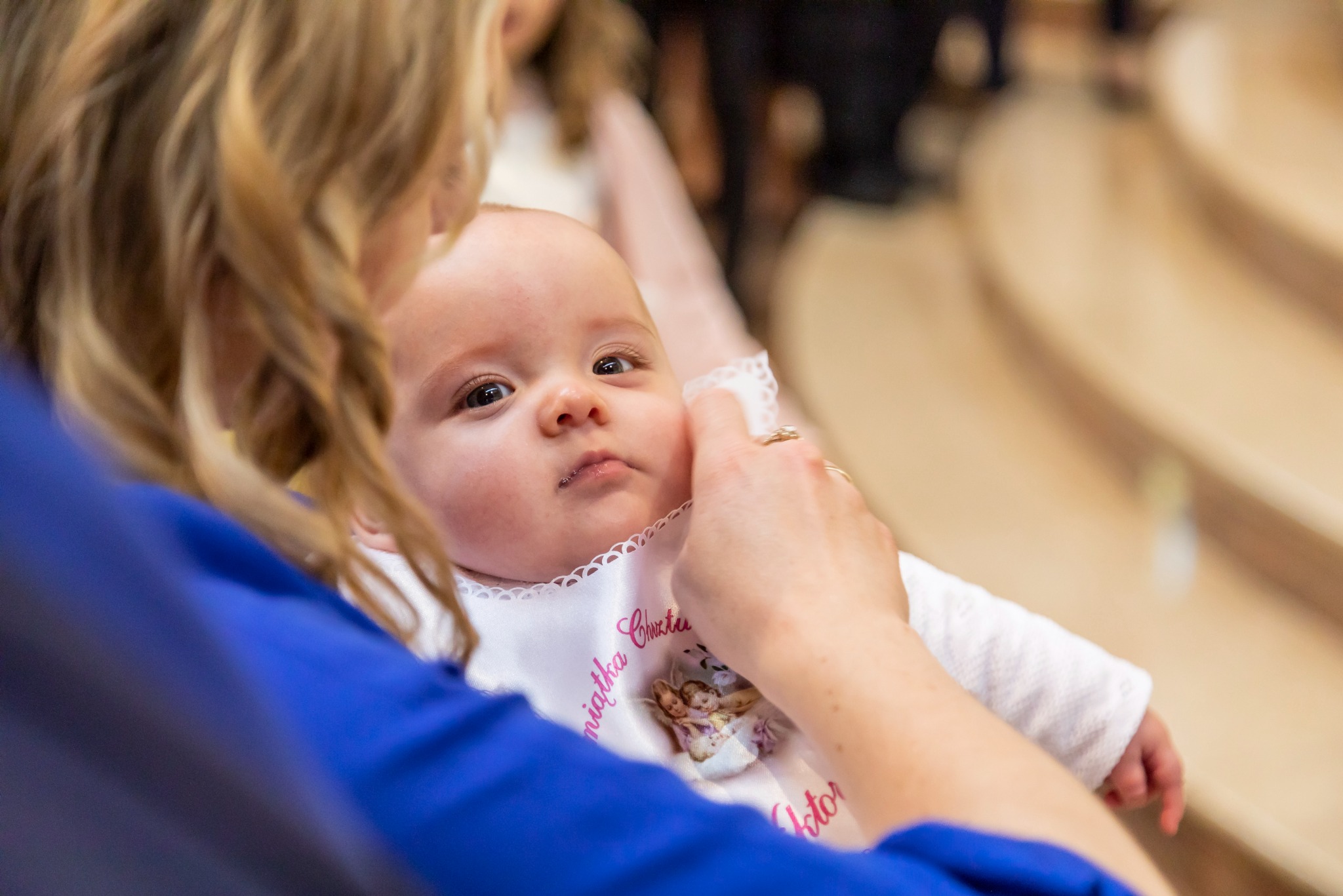 Ujęcie z góry na niemowlę w białym ubranku i ozdobnym śliniaczku z napisem 'Pamiątka Chrztu' trzymane przez osobę w niebieskiej bluzce, widoczne dłonie z pierścionkiem, w tle jasne schody kościelne.