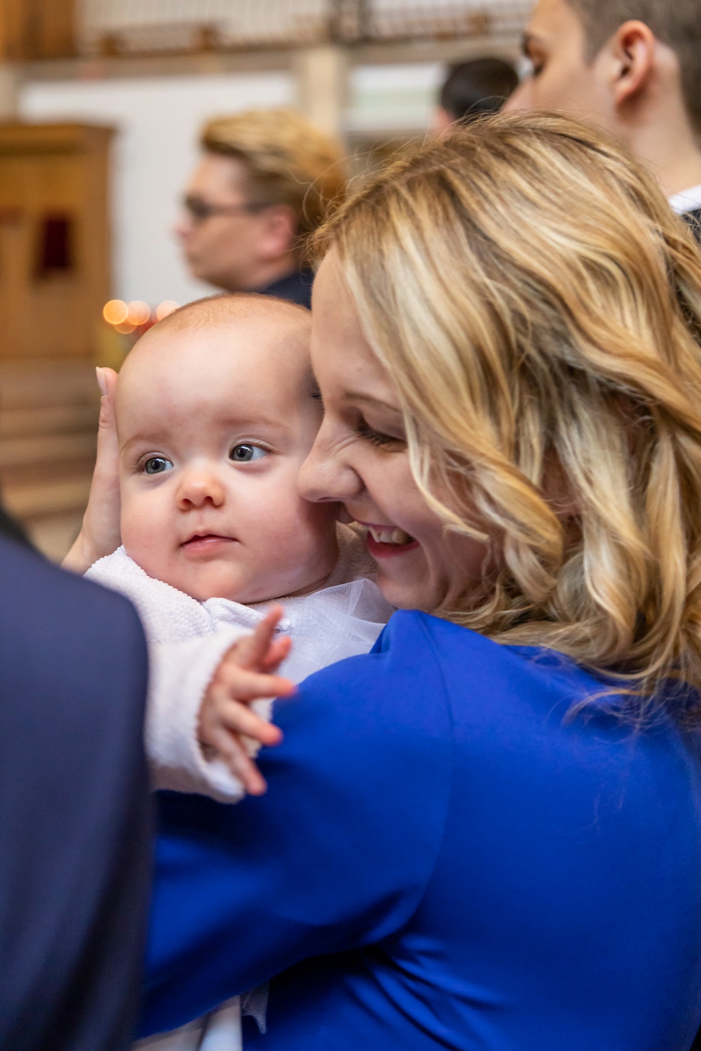 Uśmiechnięta blondynka w niebieskiej sukience trzyma na rękach niemowlę w białym ubranku, zarys wnętrza kościoła w tle.