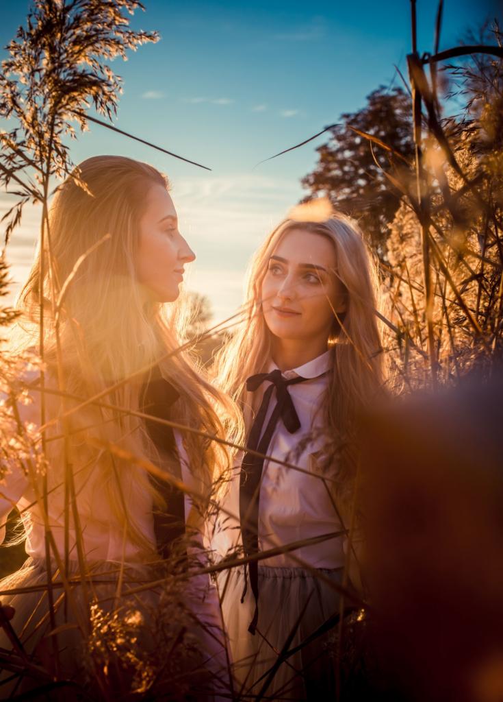 Dwie kobiety w białych bluzkach z czarnymi kokardami, stojące w wysokiej trawie w promieniach zachodzącego słońca, z delikatnym uśmiechem na twarzach.