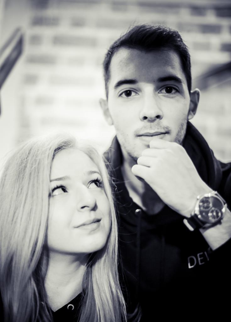 Portret czarno-biały młodej kobiety z blond włosami spoglądającej w górę i młodego mężczyzny z zegarkiem na ręce, w tle ceglana ściana.