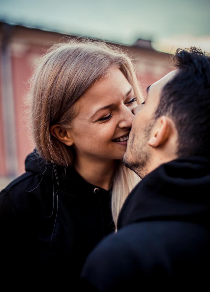 Portret uśmiechniętej, jasnowłosej kobiety w czarnej bluzie, całującej się z brodatym mężczyzną w czarnej bluzie na tle miejskiej zabudowy.