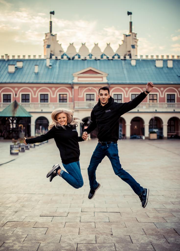 Uśmiechnięta para w czarnych bluzach i jeansach skacze trzymając się za ręce na tle Zamku Lubelskiego, oboje w ruchu, dynamiczne ujęcie.