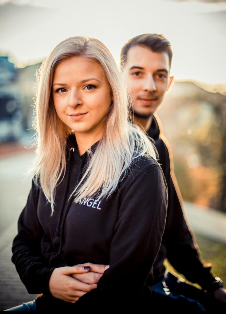 Portret młodej kobiety z blond włosami w czarnej bluzie z kapturem z napisem 'ANGEL', za nią lekko rozmyty mężczyzna, oboje na tle miejskiego krajobrazu w ciepłym świetle zachodzącego słońca.