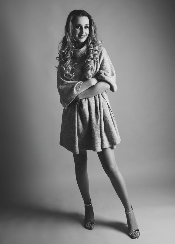 Portret w czerni i bieli: Uśmiechnięta kobieta z długimi, kręconymi włosami, ubrana w dzianinową sukienkę z szerokimi rękawami i sandały na obcasie, pozuje na jednolitym, jasnoszarym tle.