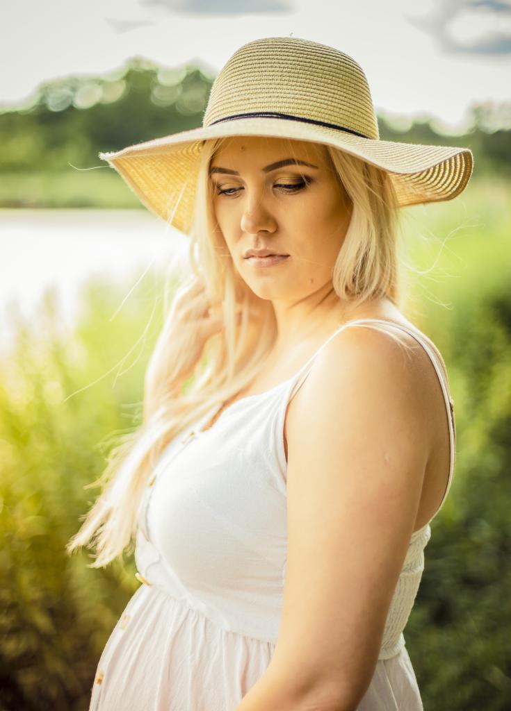 Portret kobiety w ciąży w białej sukience i słomkowym kapeluszu na tle zieleni i jeziora, z delikatnym oświetleniem i subtelnym wyrazem twarzy.