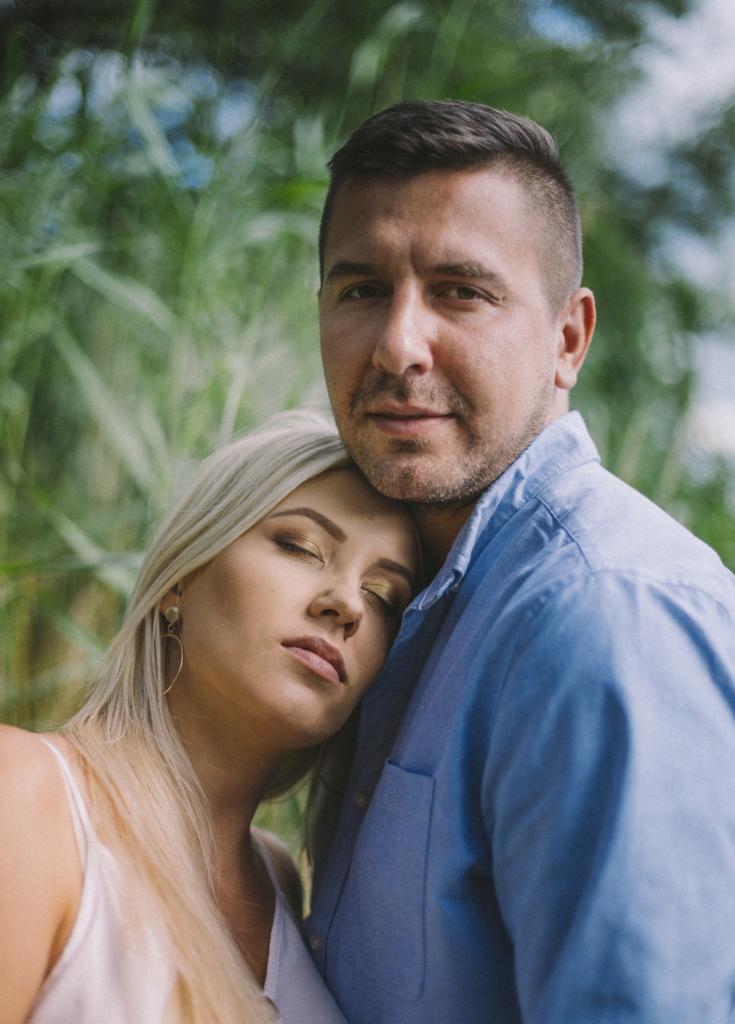 Portret pary na tle zieleni: blondwłosa kobieta z zamkniętymi oczami opiera głowę na ramieniu mężczyzny w niebieskiej koszuli.