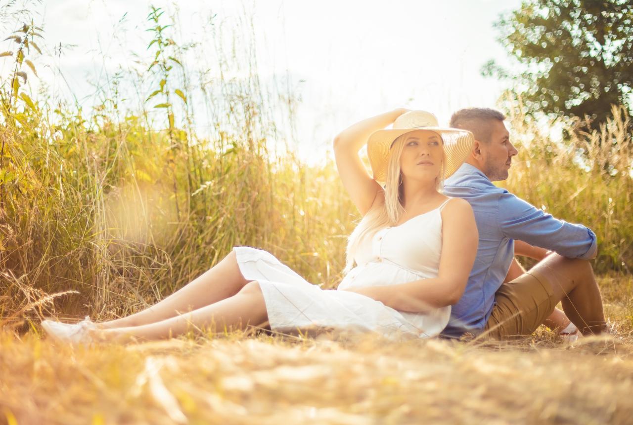 Ciężarna kobieta w białej sukience i kapeluszu siedzi w polu, opierając się plecami o mężczyznę w błękitnej koszuli, ciepłe światło słoneczne.