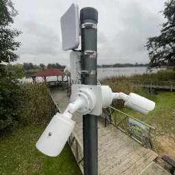 Słup z zamontowanymi trzema białymi kamerami monitoringu i anteną WiFi na tle jeziora i drewnianej kładki.