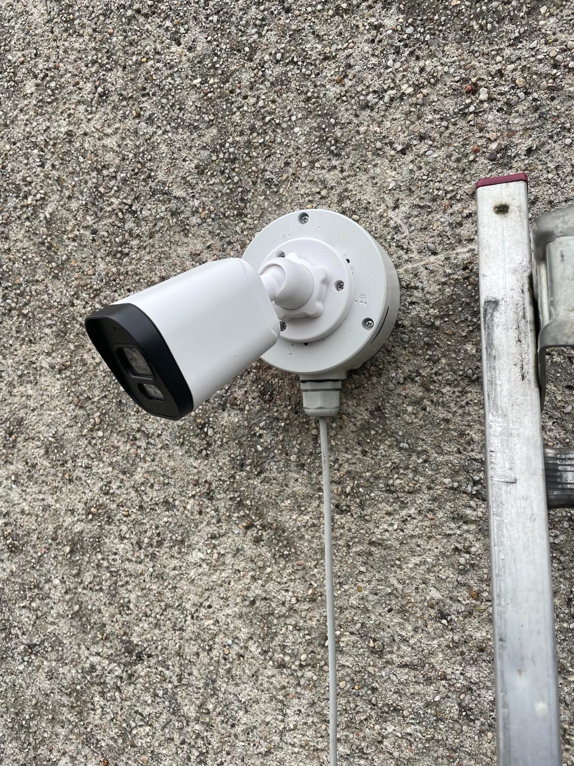 Biała kamera monitoringu z czarnym daszkiem zamocowana na szorstkiej, kamiennej ścianie, obok drabina.