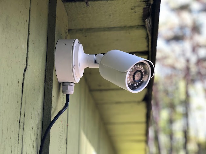 Biała kamera monitoringu zamontowana na pomalowanej na zielono drewnianej ścianie budynku, widoczny kabel zasilający.