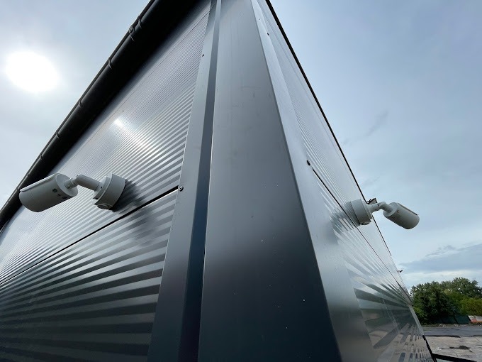 Dwie białe kamery monitoringu zamontowane na szarej, blaszanej ścianie budynku przemysłowego, widok pod kątem, pochmurne niebo w tle.