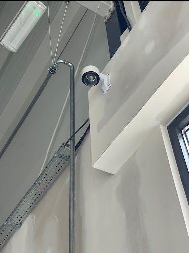 Kamera monitoringu przymocowana do ściany wewnątrz budynku, widoczne rury i kable, surowy stan wykończenia wnętrza.