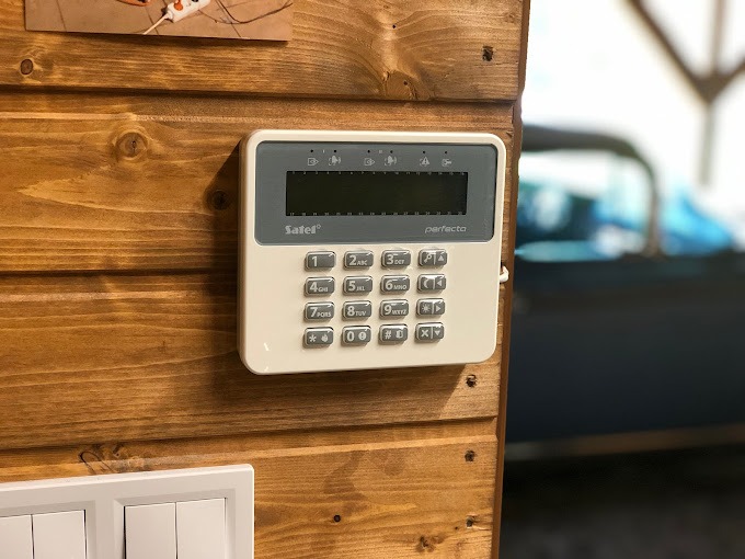 Panel sterowania alarmem Satel Perfecta zamontowany na drewnianej ścianie, widoczne przyciski numeryczne i wyświetlacz, w tle zamazany fragment niebieskiego pojazdu.