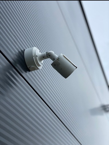 Zewnętrzna kamera monitoringu, biała, zamontowana na szarej, ryflowanej ścianie budynku, skierowana w dół pod kątem.