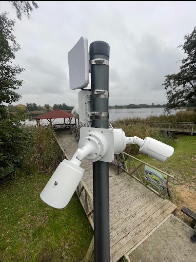 Słup z zamontowanymi trzema białymi kamerami monitoringu i anteną WiFi na tle jeziora i drewnianej kładki.