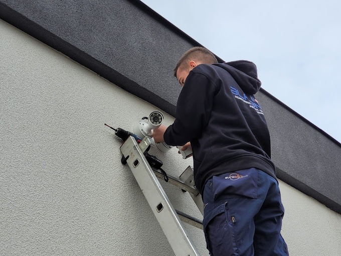 Instalator na drabinie montuje kamerę monitoringu na elewacji budynku, widoczny wkrętak akumulatorowy i logo firmy na bluzie montera.