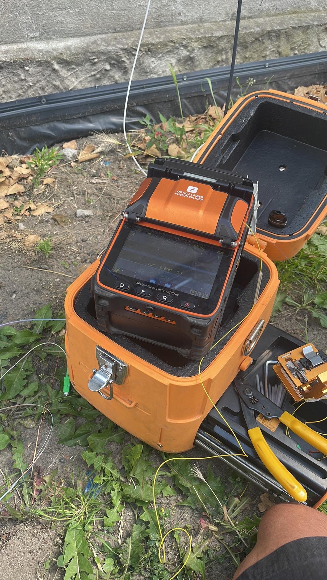 Pomarańczowa spawarka światłowodowa Optical Fiber Fusion Splicer w otwartej walizce, obok narzędzia do obróbki kabli światłowodowych, na tle trawy i betonowego muru.