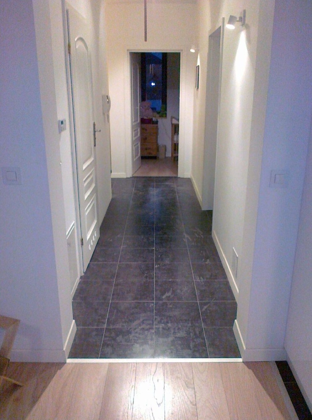 Korytarz z ciemnymi, kwadratowymi płytkami na podłodze, przechodzącymi w jasną drewnianą podłogę. Białe ściany z listwami przypodłogowymi i oświetleniem punktowym. Widoczne drzwi i fragment mebli...