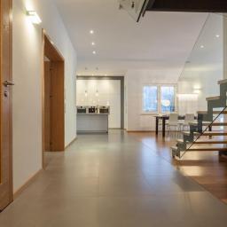 Kinvest - Nowoczesny przedpokój z szarą podłogą, drewnianymi drzwiami i balustradą szklaną przy schodach, widok na jasną kuchnię i jadalnię w głębi.