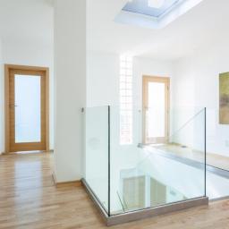 Kinvest - Jasne wnętrze z drewnianą podłogą i szklaną balustradą na antresoli, doświetlone oknem dachowym i dekorowane abstrakcyjnym obrazem na ścianie.