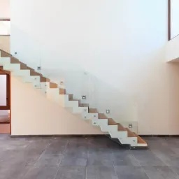 Nowoczesne schody z drewnianymi stopniami, szklaną balustradą mocowaną punktowo, oświetleniem LED w ścianie i szarą posadzką z płytek.