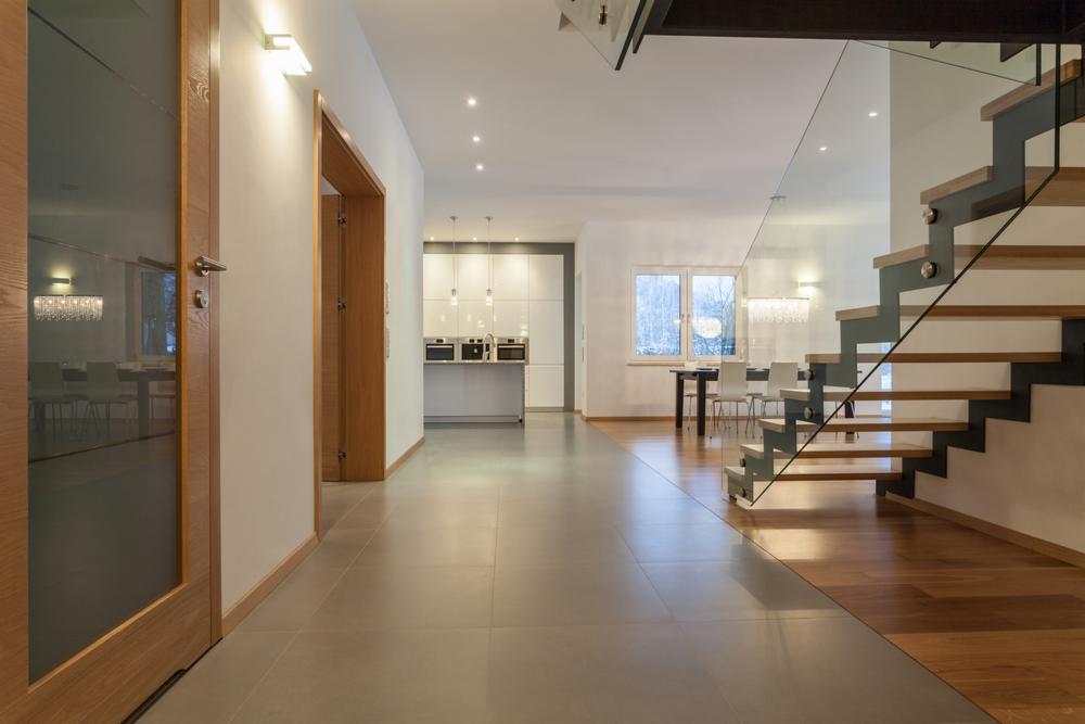 Nowoczesny przedpokój z szarą podłogą, drewnianymi drzwiami i balustradą szklaną przy schodach, widok na jasną kuchnię i jadalnię w głębi.