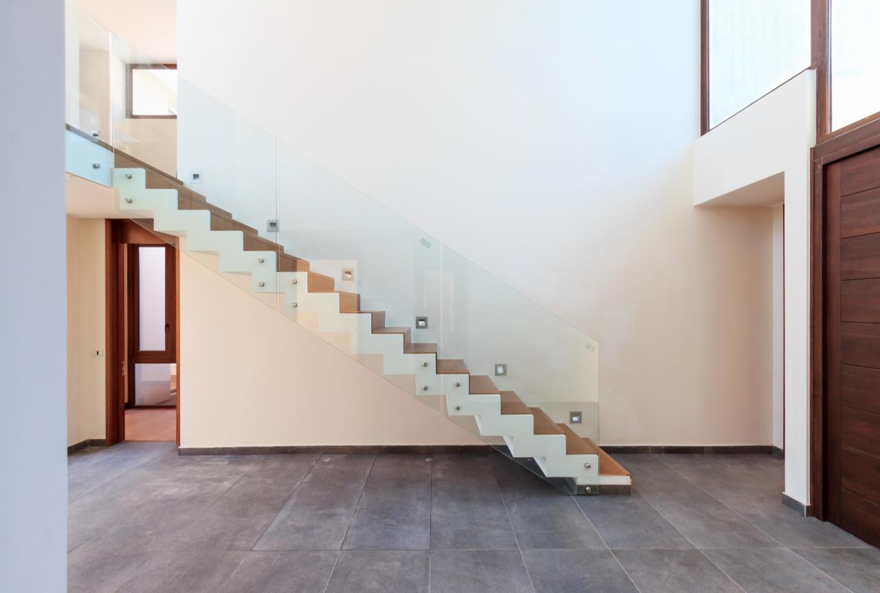 Nowoczesne schody z drewnianymi stopniami, szklaną balustradą mocowaną punktowo, oświetleniem LED w ścianie i szarą posadzką z płytek.