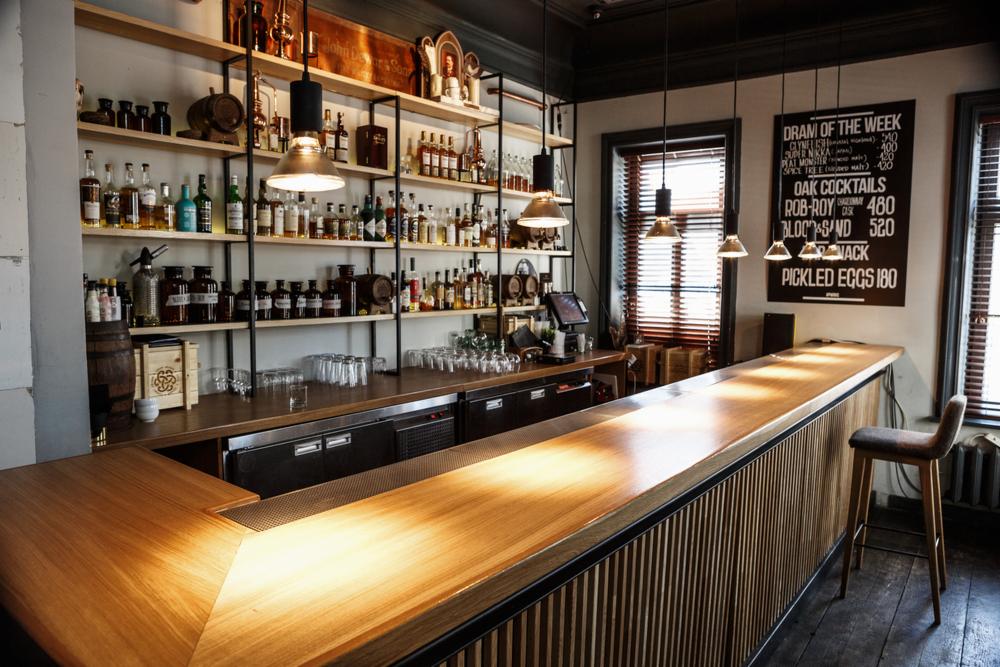 Nowoczesny bar z drewnianym blatem i frontem z pionowych listew, oświetlony wiszącymi lampami, z półkami pełnymi butelek alkoholi w tle i tablicą z menu drinków