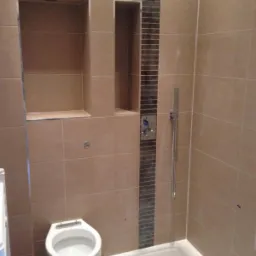 Beżowa łazienka z kabiną prysznicową typu walk-in, toaletą kompaktową i wnękami w ścianie wyłożonymi płytkami, z dodatkowym paskiem dekoracyjnym z ciemnych płytek przy natrysku.