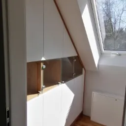 Biała szafa wnękowa na poddaszu ze szklanymi frontami, widoczne drewniane wnętrze półek, drewniana podłoga, okno dachowe z widokiem na drzewa.