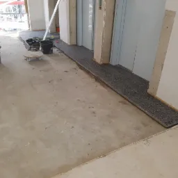 Przygotowanie podłoża pod posadzkę w przestrzeni publicznej, widoczne narzędzia i materiały budowlane, fragment ułożonej posadzki z ciemnego kamienia przy windzie.