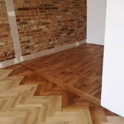 Fragment podłogi z ułożonym parkietem w jodełkę w dwóch odcieniach drewna, graniczący z czerwoną ceglaną ścianą i białym narożnikiem, widoczne ślady po remoncie.