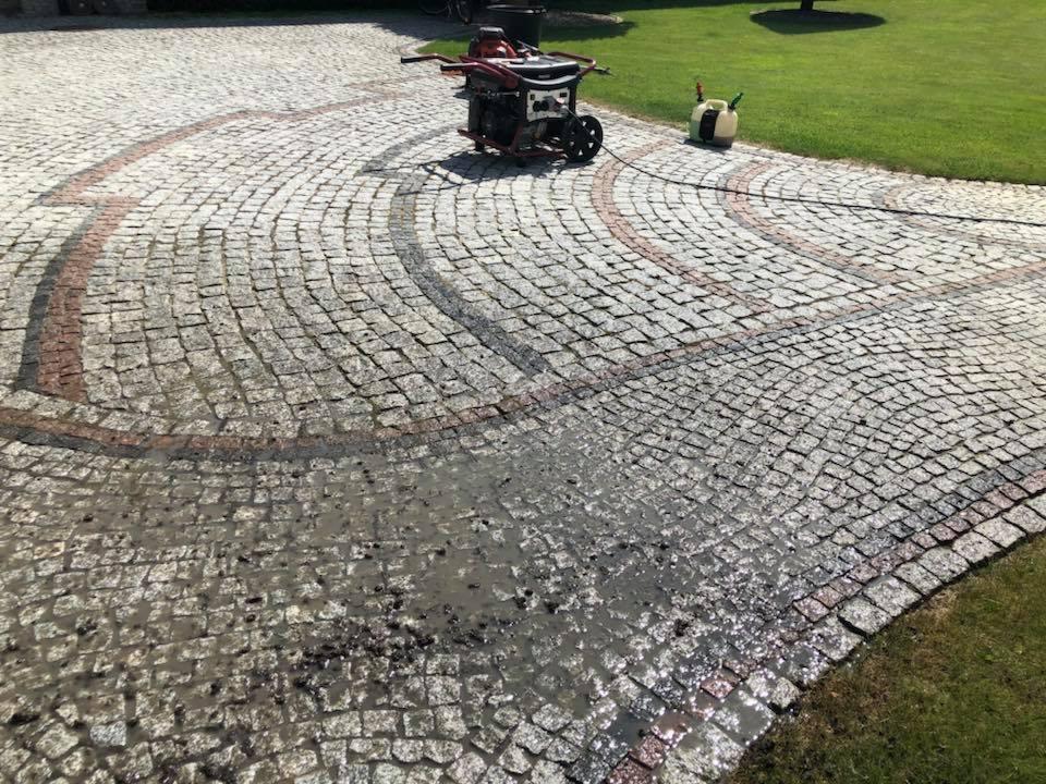 Czyszczenie kostki brukowej z użyciem myjki ciśnieniowej, widoczne zabrudzenia i woda na powierzchni, wzór z czerwonej kostki.