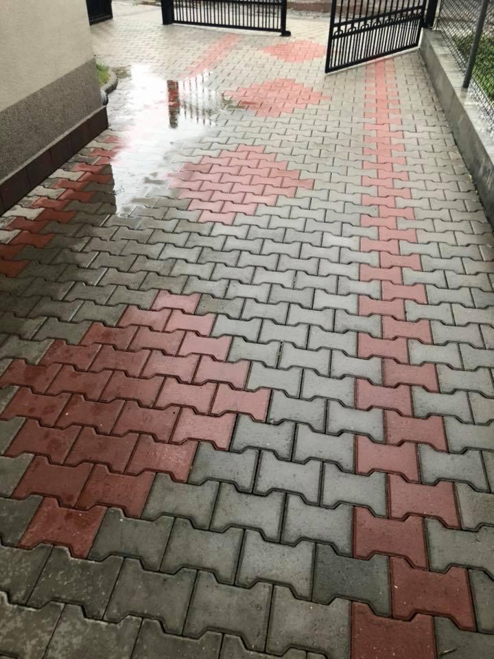 Mokry chodnik z kostki brukowej w odcieniach szarości i czerwieni, z widocznymi śladami wody po czyszczeniu, brama w tle.