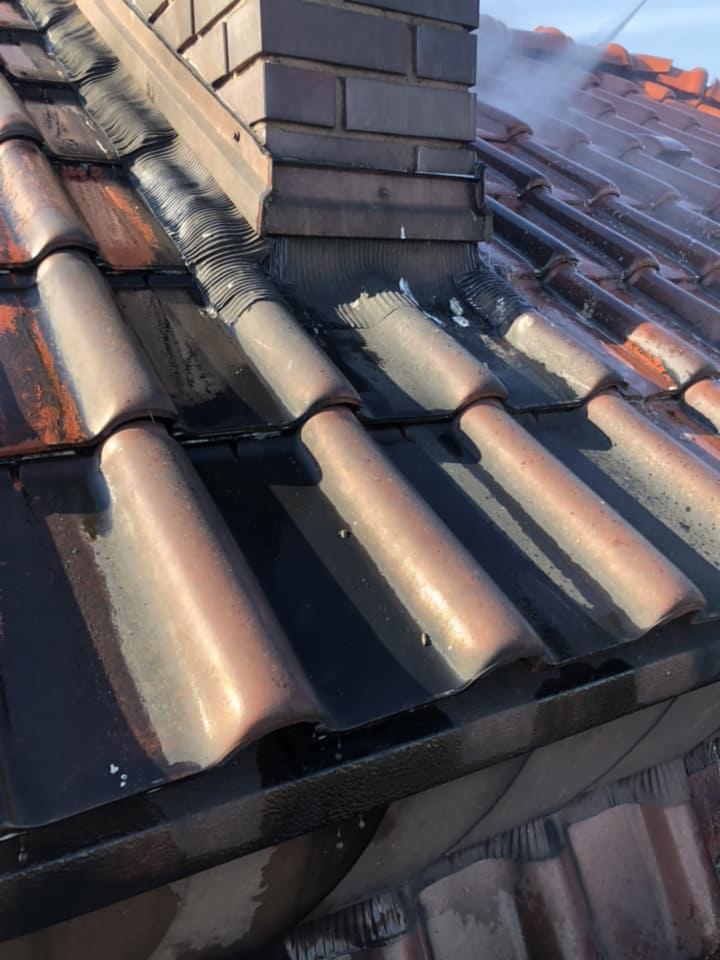 Czyszczenie dachu z ceglanej dachówki przy kominie za pomocą myjki ciśnieniowej, widoczna para wodna i mokra powierzchnia.