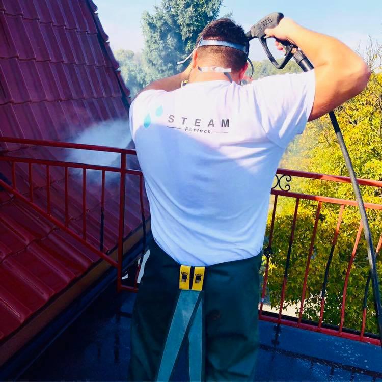 Pracownik w białej koszulce z logo firmy czyści czerwoną dachówkę za pomocą myjki ciśnieniowej, widoczne drzewa w tle.