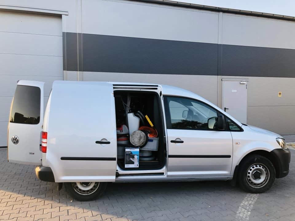 Srebrny van z otwartymi drzwiami bocznymi i tylnymi, widoczne wyposażenie do czyszczenia wewnątrz, zaparkowany na kostce brukowej przed szarym budynkiem.