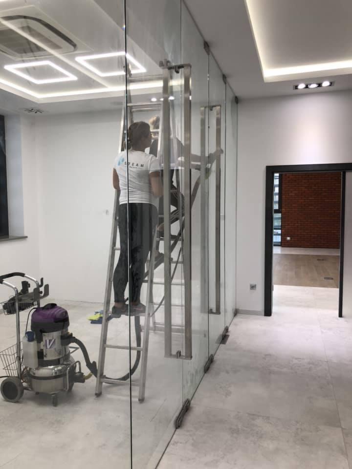 Pracownik na drabinie czyści szklane ściany biurowe za pomocą profesjonalnego odkurzacza do wody, widoczne uchwyty i mocowania szkła.