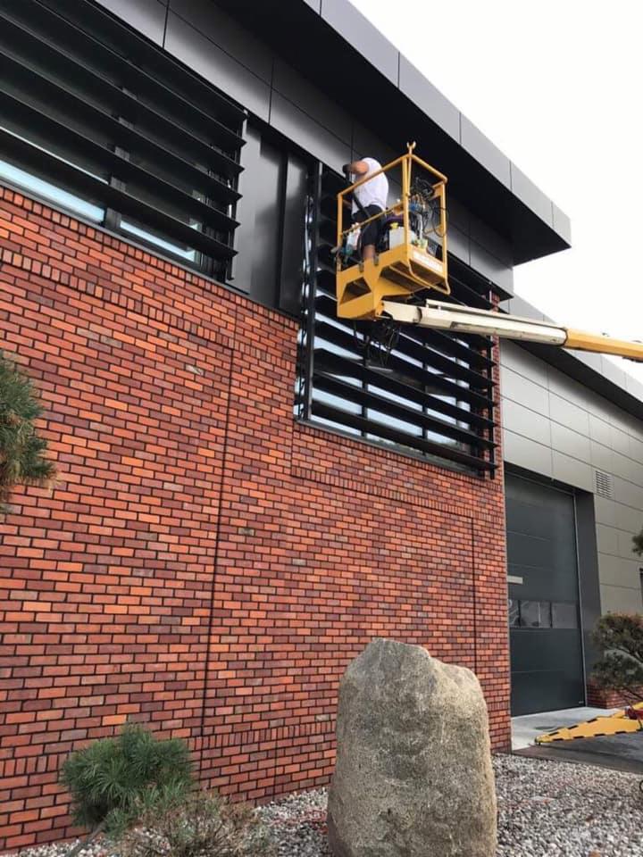 Pracownik w podnośniku koszowym czyści okna biurowca z ceglaną elewacją i czarnymi żaluzjami.