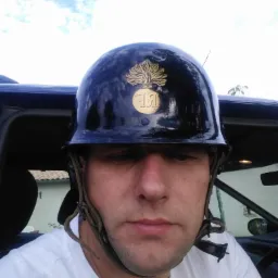 Mężczyzna w hełmie z emblematem, siedzący w samochodzie, z widocznym niebem w tle. Hełm jest czarny, błyszczący, z motywem roślinnym.