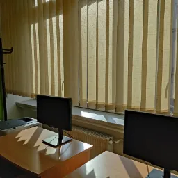 Sala komputerowa z dwoma stanowiskami pracy, każde z monitorem LCD, biurkiem z blatem w kolorze jasnego drewna, na ścianie zielona farba, okno zasłonięte pionowymi żaluzjami w odcieniach beżu,...