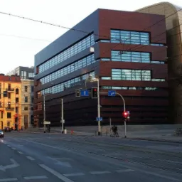 Nowoczesny budynek biurowy z brązową elewacją i rzędami okien, widoczny z perspektywy ulicy z tramwajowymi torami i sygnalizacją świetlną.