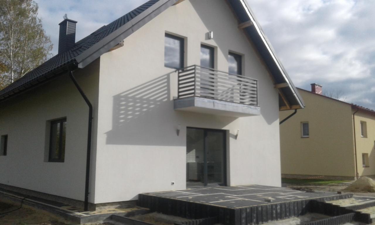 Nowoczesny dom jednorodzinny z balkonem z metalową balustradą o poziomych szczeblach, tarasem wyłożonym płytami i czarną dachówką, w trakcie prac wykończeniowych na zewnątrz.