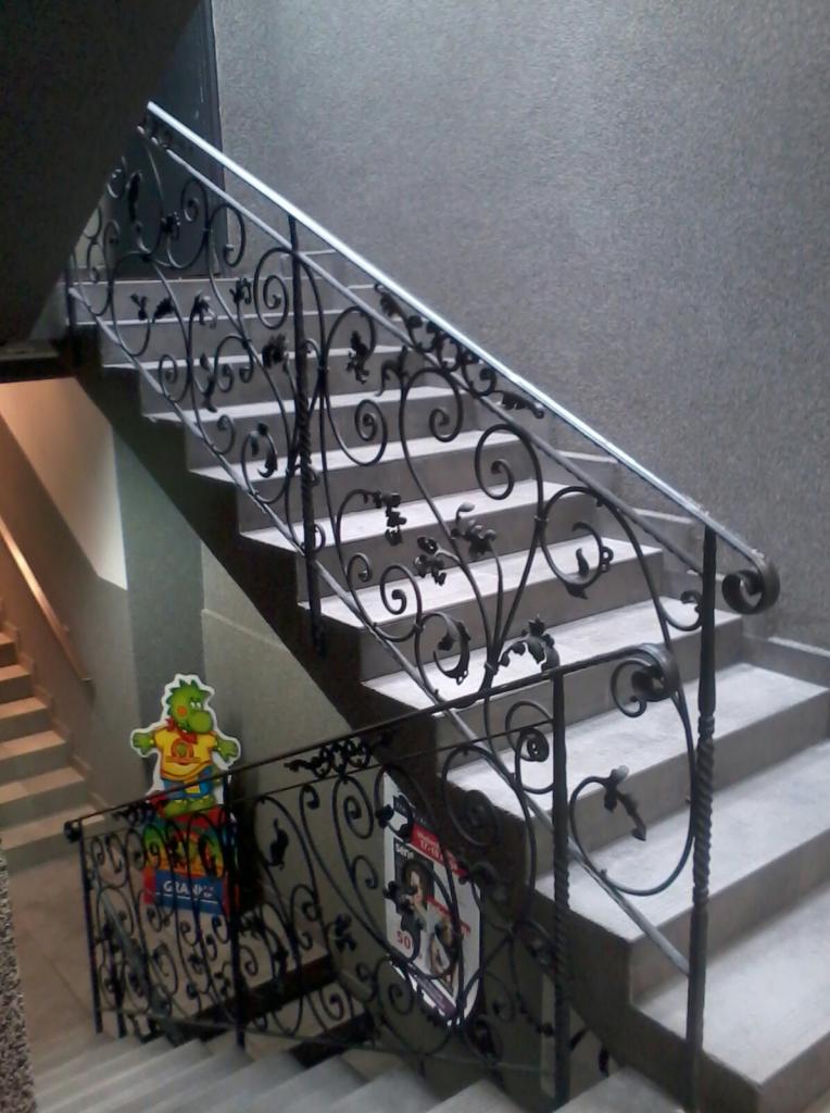 Kuta balustrada schodowa z motywem roślinnym, widoczna z dołu, z plakatem i kartonową figurką na pierwszym planie, szare ściany i schody.