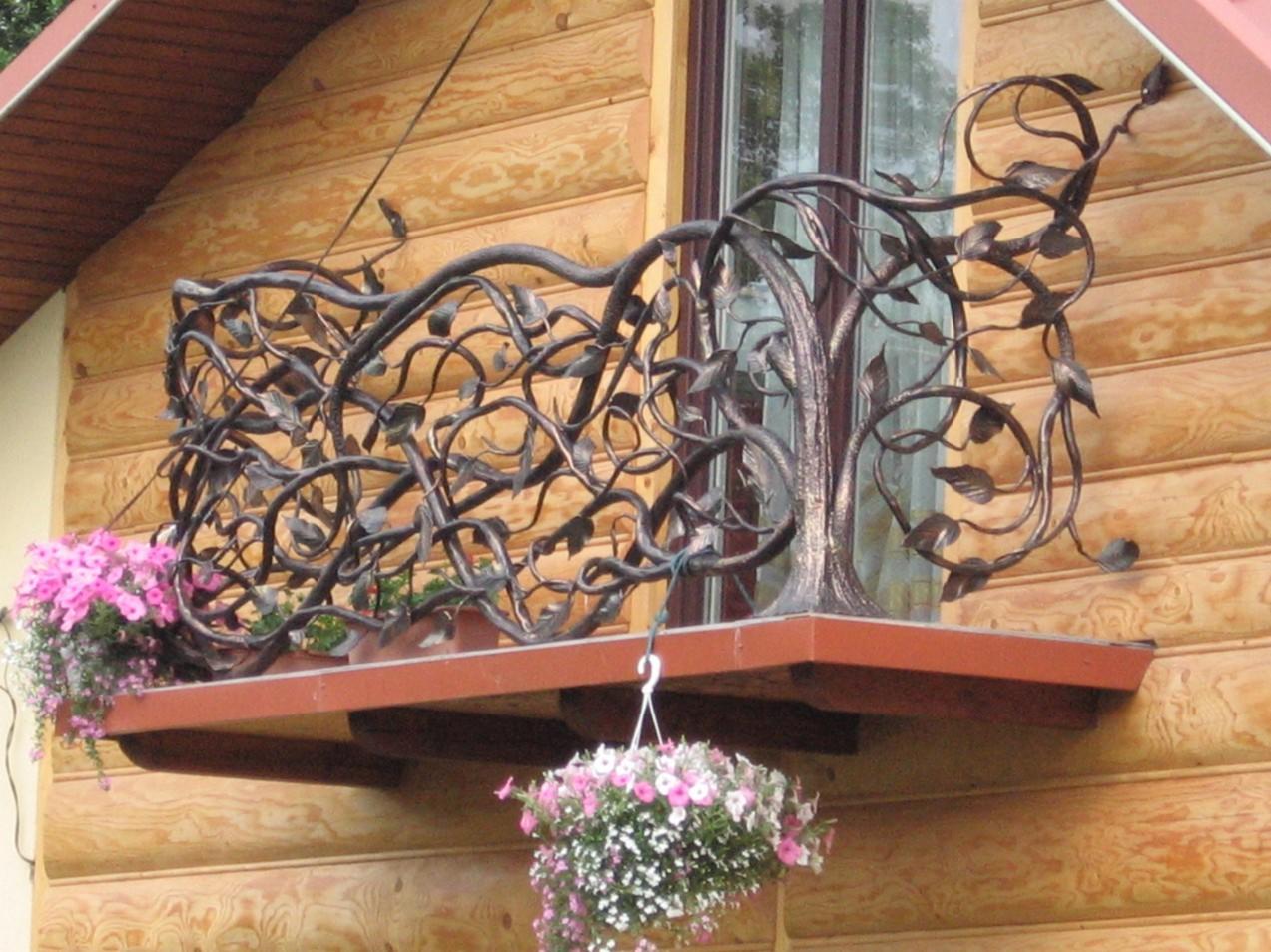 Kuta balustrada balkonowa z motywem drzewa, ozdobiona kwiatami w doniczkach i wiszącą kompozycją kwiatową, na tle drewnianej elewacji domu.