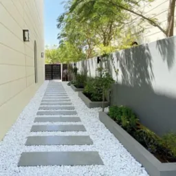 Ogród z ścieżką z betonowych płyt, wysypaną białym żwirem, z minimalistycznymi rabatami wzdłuż szarego muru.