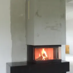 Nowoczesny kominek narożny z czarną obudową i betonową konstrukcją w surowym wnętrzu, widoczne płomienie i niedokończone ściany.