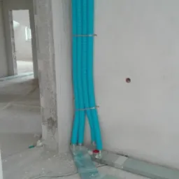 Turkusowe, karbowane rury wentylacyjne przymocowane do ściany w surowym wnętrzu, z widocznymi kanałami wentylacyjnymi na podłodze, ukazujące etap instalacji.